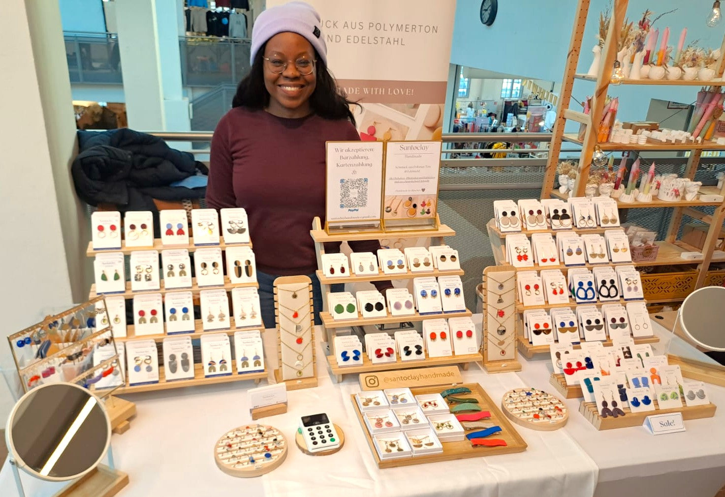 Flávia Troppmann, junge Schmuckdesignerin präsentiert handgemachte Ohrringe, Ketten und Accessoires von Santoclay Handmade an einem liebevoll dekorierten Marktstand – ideal für moderne, farbenfrohe Schmuckliebhaber.
