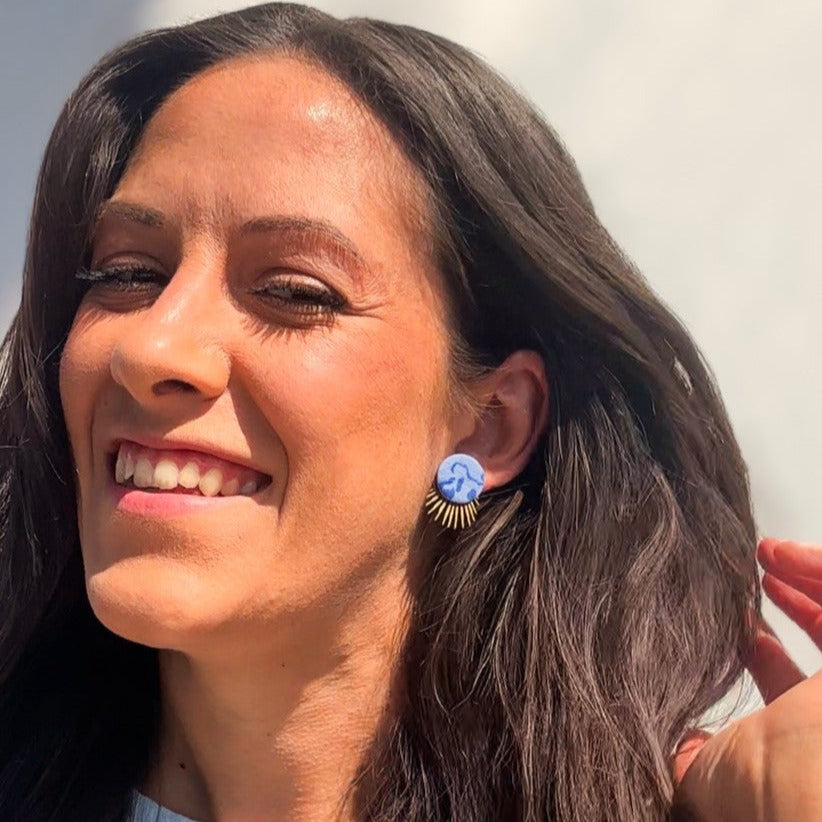 Frau mit langen braunen Haaren trägt handgemachte blaue Statement-Ohrstecker mit goldfarbenem Sonnenstrahl-Detail von Santoclay und lächelt in die Sonne.