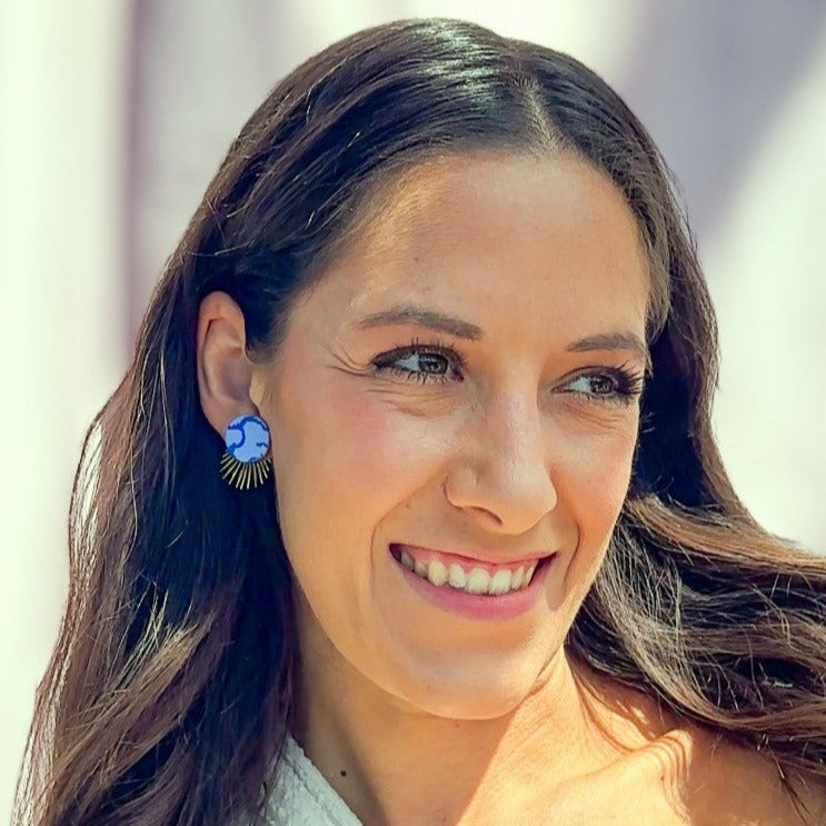 Frau mit langen braunen Haaren trägt handgemachte blaue Statement-Ohrstecker mit goldfarbenem Sonnenstrahl-Detail von Santoclay und lächelt in die Sonne.